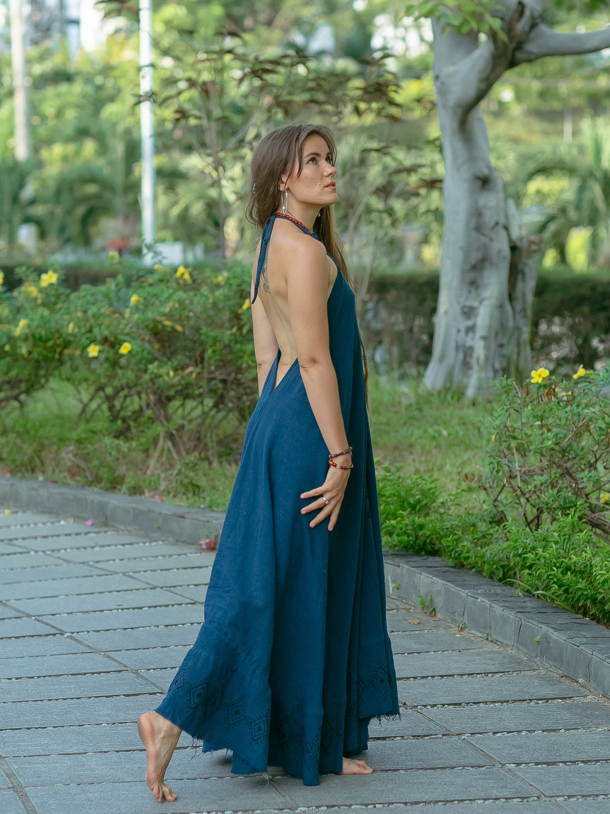 A woman wearing the Hanvi maxi dress, a long and backless style made from sustainable cotton, walks barefoot along a stone path in a garden, glancing over her shoulder with lush greenery and trees behind her.