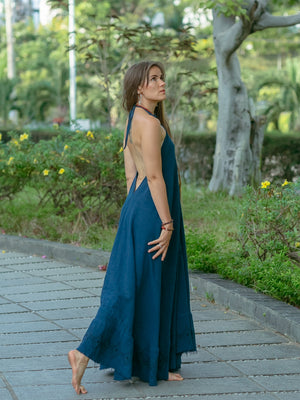 A woman wearing the Hanvi maxi dress, a long and backless style made from sustainable cotton, walks barefoot along a stone path in a garden, glancing over her shoulder with lush greenery and trees behind her.