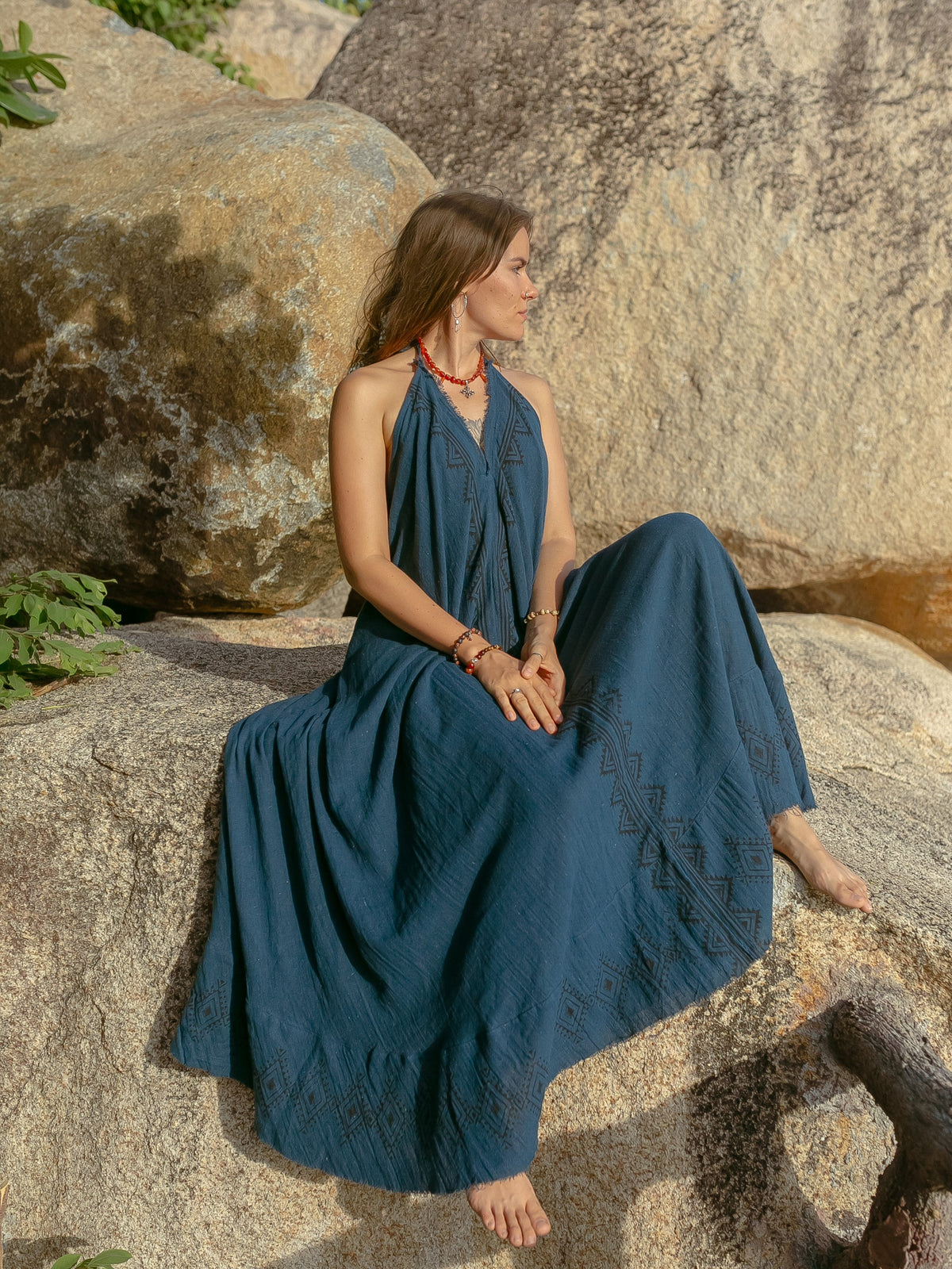 A woman in the Hanvi maxi dress, a long sleeveless blue gown made from sustainable cotton, sits barefoot on a large rock and gazes to the side. Sunlight illuminates her and the textured rocky backdrop.