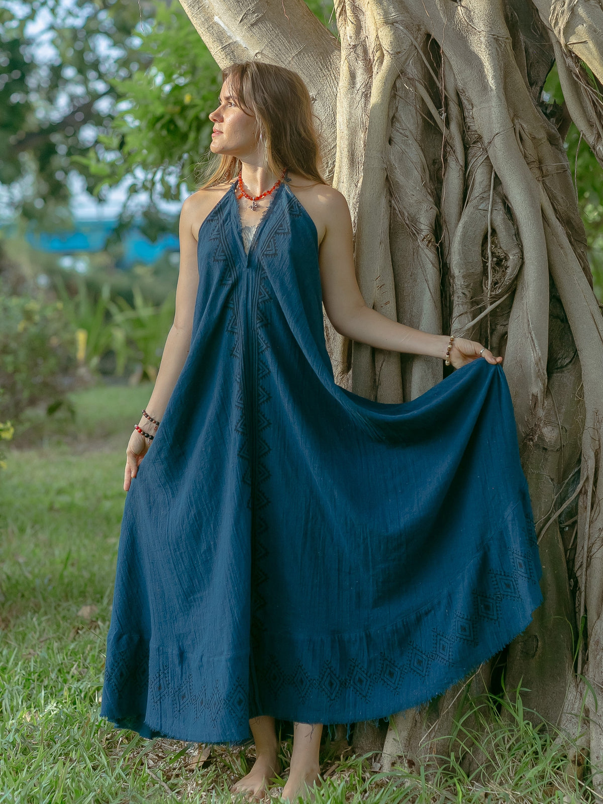 A woman stands barefoot on the grass next to a large tree with twisted roots, surrounded by greenery, wearing a blue Hanvi maxi dress made from sustainable cotton. She holds out her skirt and looks to the left.