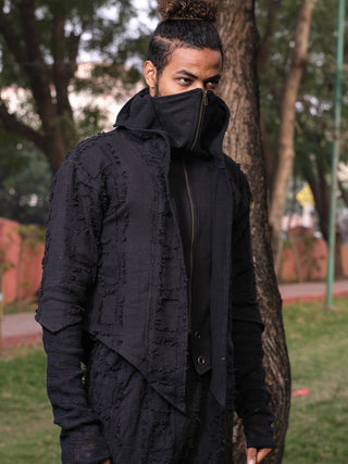Man in a black frayed cotton ninja hoodie with the stand collar zipped over his mouth, outdoors among trees