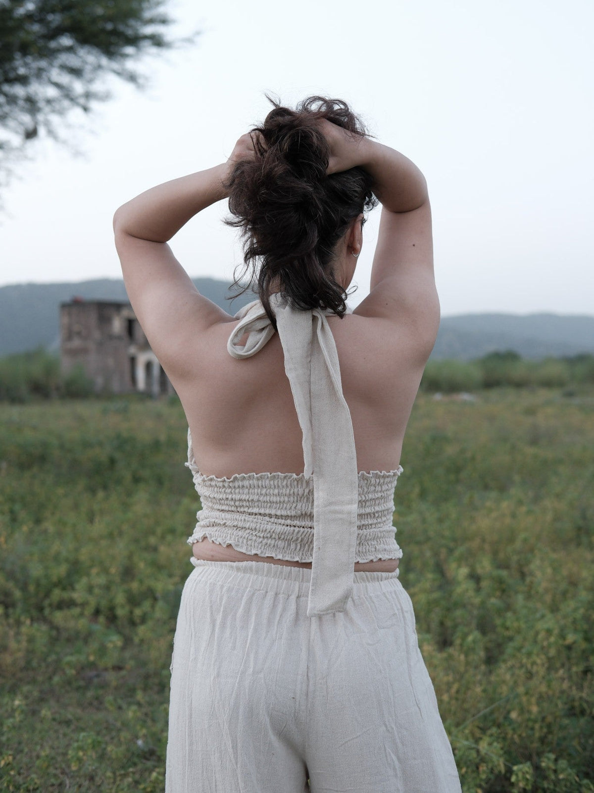 Back view of the halter top showing the open back, long self-tie knotted at the nape and smocked band, arms raised