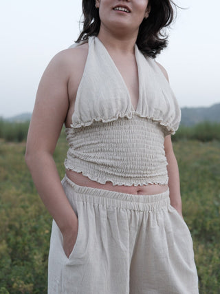 Close crop of the smocked bodice and ruffle edges on the off-white khadi halter top, hand resting in trouser pocket