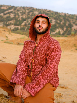 Model seated in the Rangrek hooded jacket, hood up, showing the rust geometric handblock print on a cropped cotton jacket