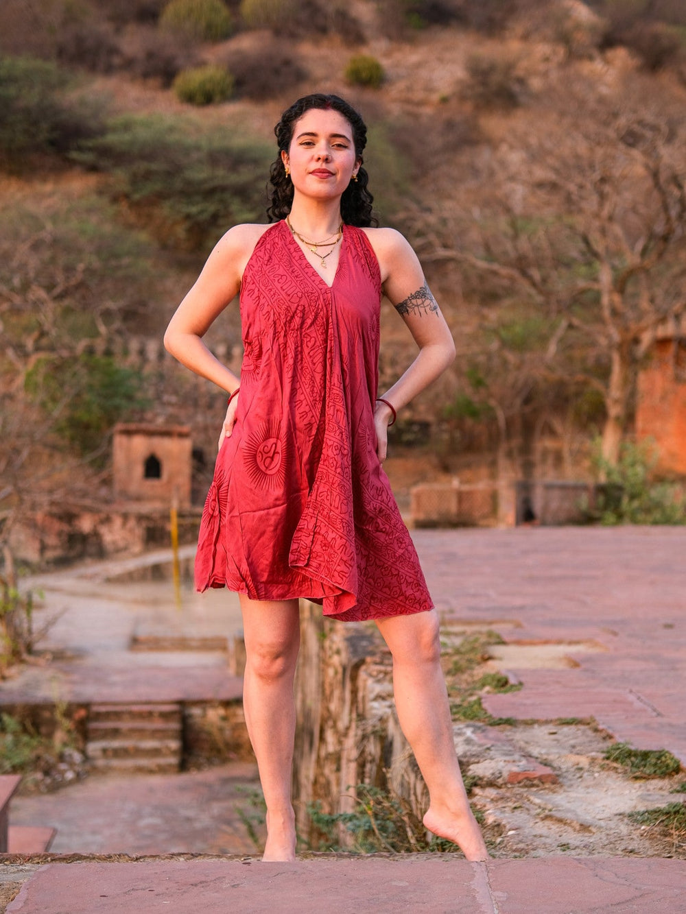 Red Om print halter cotton dress, front view with hand on hip against a heritage stone backdrop