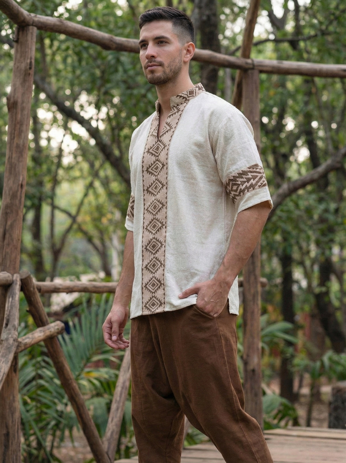 A man stands outdoors surrounded by wooden structures and lush greenery, wearing the Aho Kurta in cream, which features geometric patterns inspired by Rajasthani handblock prints, paired with brown pants.