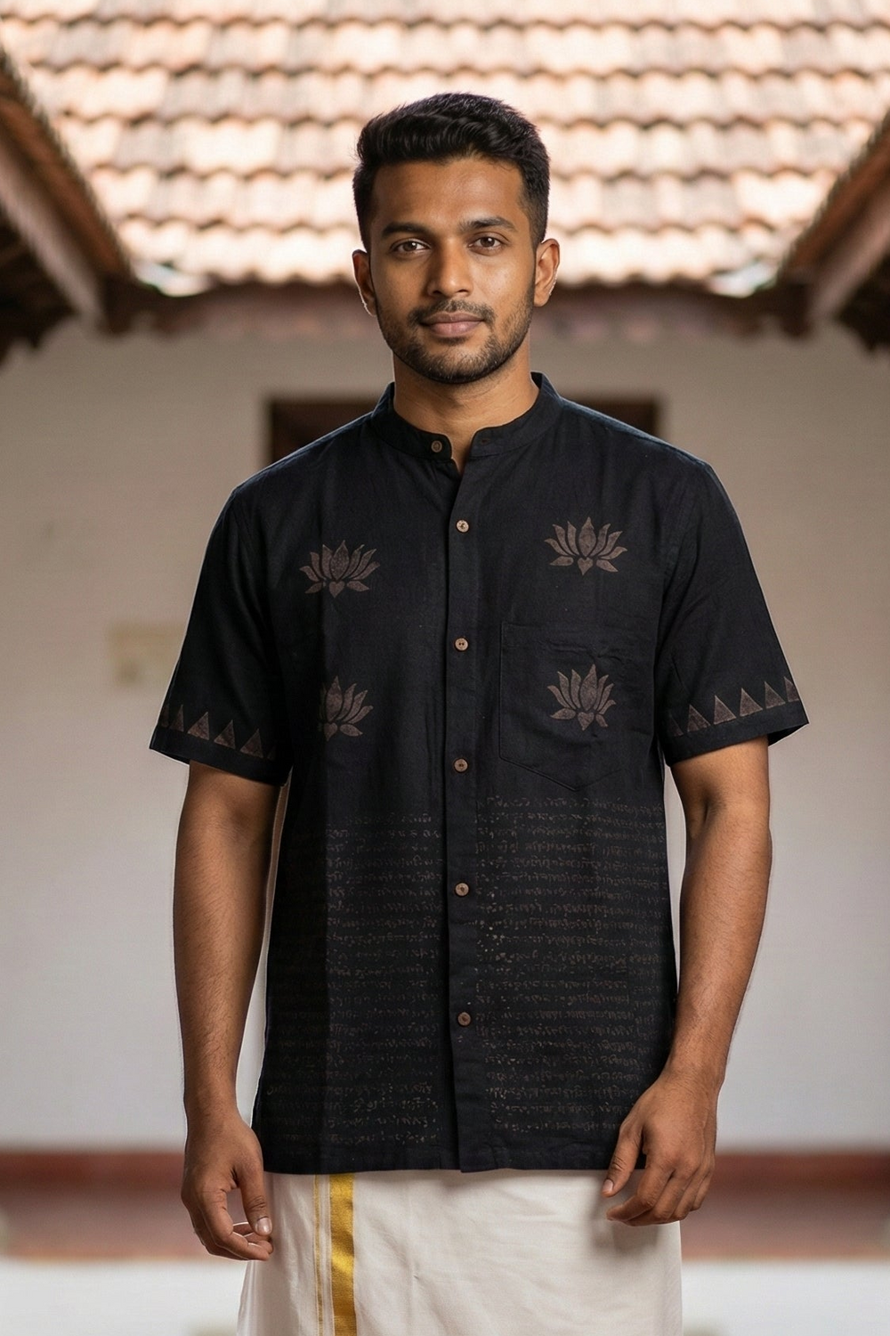 A man stands facing forward outdoors between tiled-roof buildings, dressed in the elegant Samhita Shirt—a black short-sleeved shirt adorned with lotus motifs in a block discharge print—paired with a cream-colored traditional garment featuring a yellow stripe.