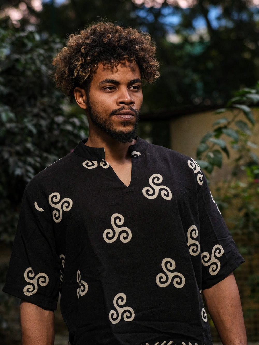 A man with curly hair and a beard wears a black Spiral Kurta, featuring a block-printed tribal spiral design, while standing outdoors. The green foliage and blurred background create a natural setting.