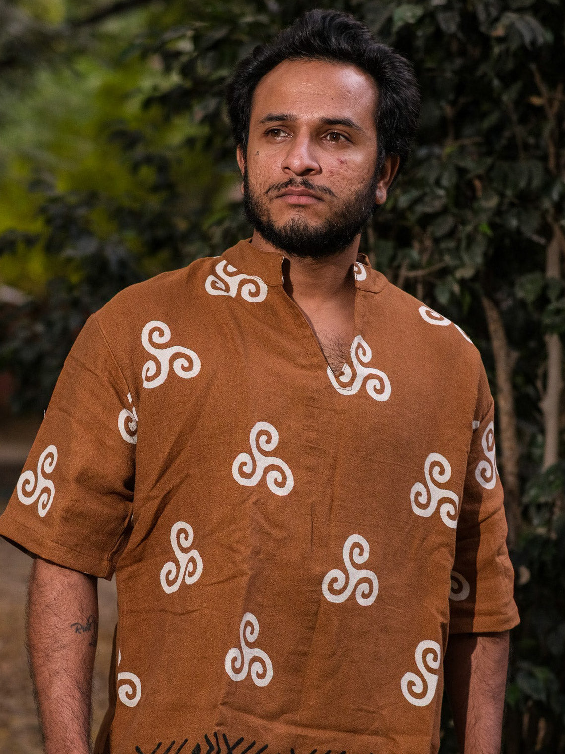 A man with a beard and short hair wears the Spiral Kurta, a brown crepe shirt with white tribal spiral designs, standing outdoors before green foliage and looking to the side with a serious expression.