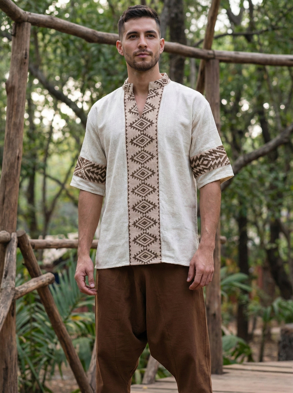 A man stands outdoors wearing the Aho Kurta, featuring natural-colored fabric embellished with brown geometric embroidery inspired by Rajasthani handblock print, paired with loose brown pants. Wooden railings and trees are visible in the background.