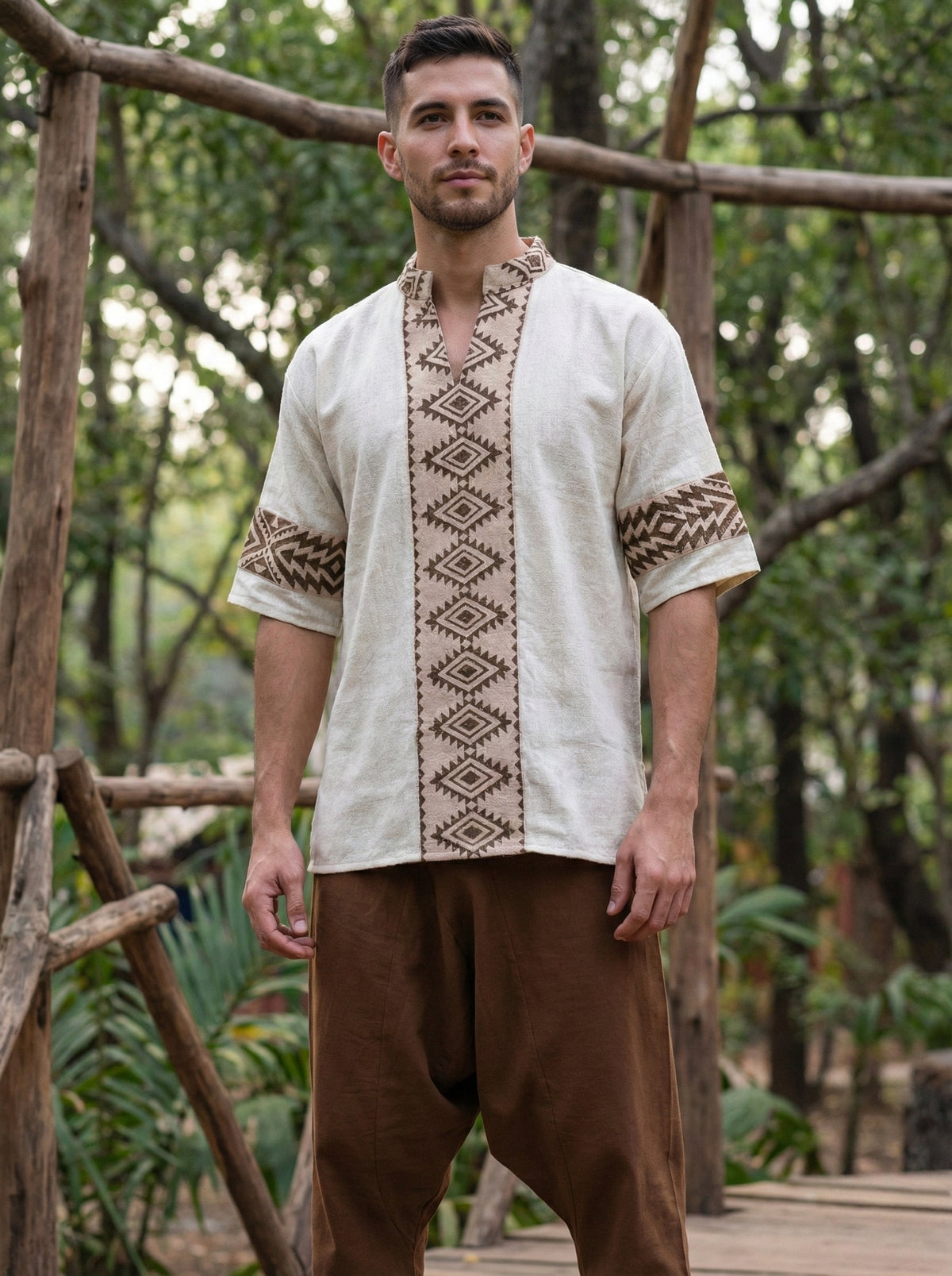 A man stands outdoors wearing the Aho Kurta, featuring natural-colored fabric embellished with brown geometric embroidery inspired by Rajasthani handblock print, paired with loose brown pants. Wooden railings and trees are visible in the background.