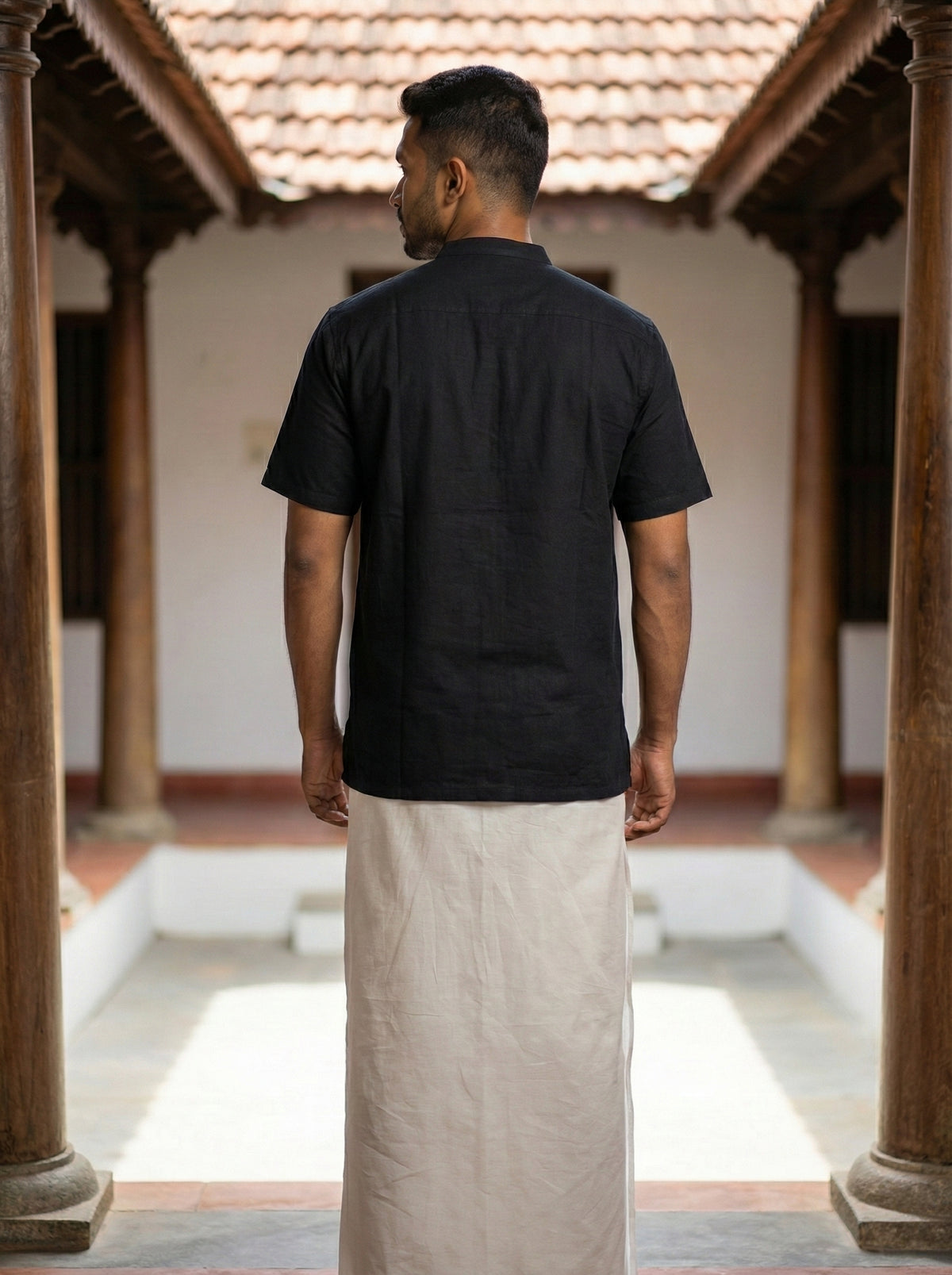 A person stands with their back to the camera, dressed in an elegant black Samhita Shirt paired with a traditional white dhoti, in a courtyard featuring wooden pillars and a tiled roof.