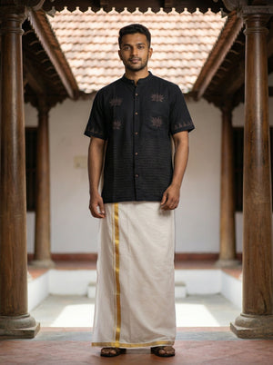 A man stands in a traditional courtyard wearing the Samhita Shirt paired with a white mundu featuring a gold border, looking at the camera.