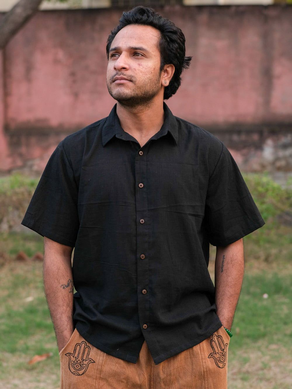 A man wearing the black Yantra Shirt made from breathable cotton and tan pants stands outdoors with his hands in his pockets, looking thoughtful. A blurred wall and grass can be seen in the background.
