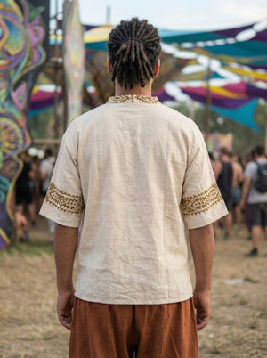 A person with short dreadlocks, dressed in the Aho Kurta—a beige shirt featuring Rajasthani handblock print trim—and brown pants, stands outdoors at a colorful festival, facing away from the camera. Other people and vibrant decorations are visible in the background.
