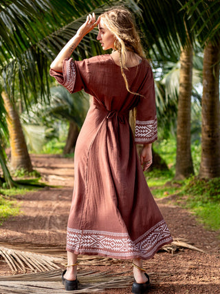 Samudra Maxi Dress brown cotton jute, model walking a shaded palm grove path, hand to hair