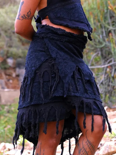 Outdoors amidst greenery, a person showcases the Titania Wrap Skirt in black, its layered fabric and frayed edges adding style. Their midriff and tattoos on their arm and leg are visible in the image.