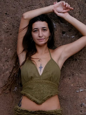 A woman with long brown hair poses against a textured brown wall, wearing an olive green Titania Crop Top and necklace, her arm raised above her head and a gentle expression on her face.