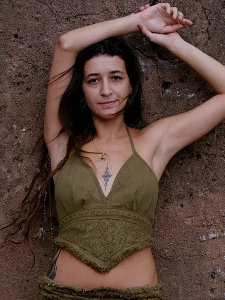A woman with long brown hair poses against a textured brown wall, wearing an olive green Titania Crop Top and necklace, her arm raised above her head and a gentle expression on her face.