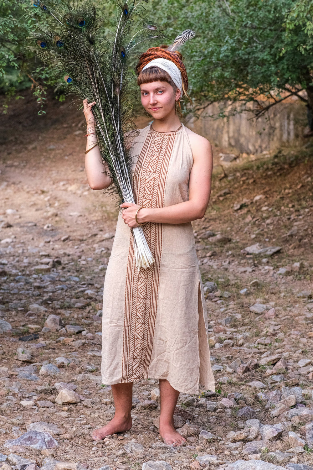 Beige Trishika khadi cotton halter maxi dress on model holding a fan of peacock feathers on a stony path