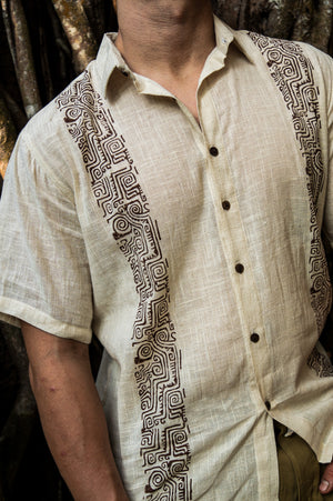 A person wearing the Utopian Shirt in natural cream—a short-sleeve shirt featuring a geometric black tribal design and dark buttons—standing in front of a textured, tree bark background.